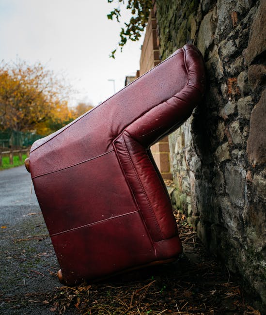 An upright maroon leather sofa is positioned outside against a stone wall on a pavement, with the backrest leaning against the wall and the base resting on the ground. The sofa's arms and cushions are visible, showing some wear and minor scratches. The surrounding environment includes autumn foliage with orange and green leaves in the background, a partly cloudy sky, and nearby residential buildings. This outdoor scene depicts the initial stages of a house removal process where furniture is being prepared for transport, with the sofa awaiting loading into a van. Man with Van Sanderstead, a relocations company, may use such images to illustrate furniture transport, home relocation, or packing and moving activities, emphasizing the careful handling of household items during the moving process.