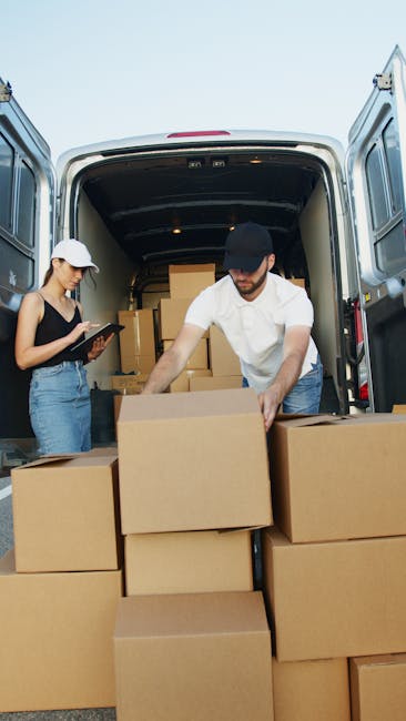 Two movers from Man with Van Sanderstead are engaged in the home relocation process, loading and organizing cardboard boxes into the back of a large commercial van on a clear day. The male mover, wearing a white shirt and dark cap, is placing a stacked box onto a pile of similar moving cartons situated at the van’s open rear doors, which reveal a spacious cargo area filled with numerous packed boxes. The female mover, dressed in a black top, jeans, and a white cap, is standing nearby holding a clipboard or tablet, likely overseeing or recording the packing process. The scene is set outdoors on a paved surface, with natural daylight illuminating the scene, reflecting a professional and systematic furniture transport and packing operation carried out by [COMPANY_NAME], enhancing the efficiency of Sanderstead's house removals services.