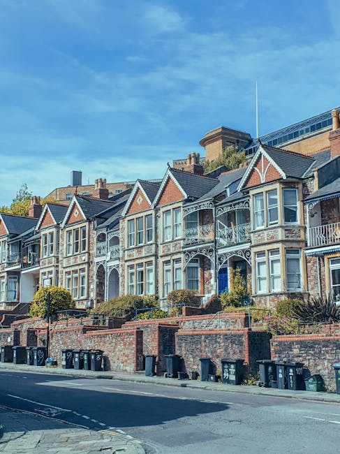 A row of Victorian terraced houses with ornate white-painted ironwork on balconies and bay windows, situated on a residential street during daytime with clear blue skies. The properties are built with red brick and have gabled roofs. In front of the houses, there is a brick retaining wall with a concrete cap, and a sidewalk lined with black wheelie bins and small shrubs. The street appears calm and clean, and the scene is associated with house moves or home relocations, reflecting the typical environment for professional removals services like those offered by Man with Van Sanderstead, especially when managing packing and furniture transport for local moves in Croydon.