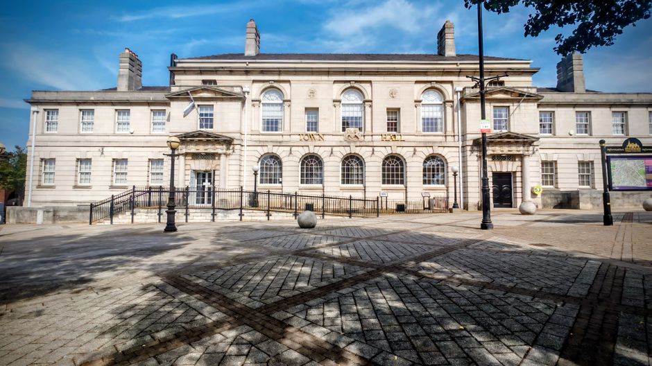A large, historic stone building with classical architectural features, including arched windows and decorative columns, situated in an outdoor urban area. The foreground shows a paved pedestrian space with a pattern of dark and light bricks, casting shadows from nearby trees and street lamps. Two large spherical stone bollards are positioned in front of the building, and a black metal railing runs along the front steps. To the right, there is a bus shelter and a lamppost, with a tree partially visible in the upper right corner. The weather appears clear with blue skies and some scattered clouds. The scene depicts a calm environment suitable for household removals, with front access likely used for furniture transport or home relocation purposes, consistent with services offered by Man with Van Sanderstead.