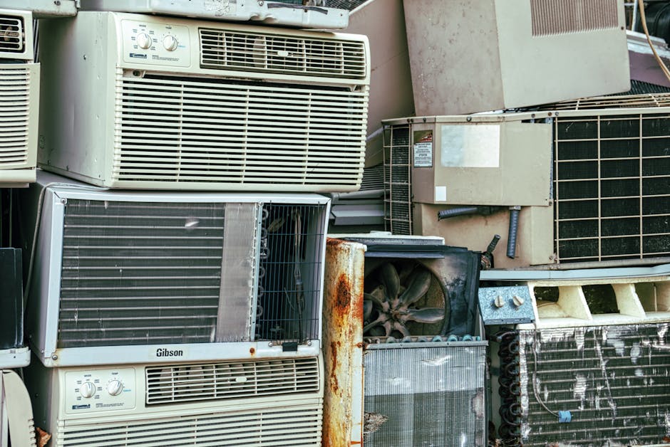 Close-up image of a piled-up collection of used air conditioning units and old radiators, stacked against a wall outside a property. The units include various sizes and models, with some showing visible rust, dirt, and damage. The pile comprises metal and plastic components, with some units featuring exposed fans and internal parts. Among the objects are items wrapped in protective materials such as cardboard and plastic. The environment appears to be an outdoor area used for storing or discarding old appliances, with the equipment arranged haphazardly. This scene reflects the type of bulk waste items that might be handled by a professional removals service like Man with Van Sanderstead during a home relocation or appliance disposal process. The image highlights the importance of proper waste management consistent with borough rules, such as those outlined by Croydon Council, for responsible appliance disposal during house moves or renovations.