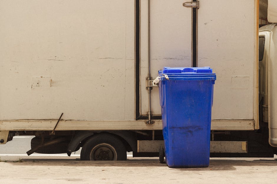 A blue recycling bin with a closed lid is positioned on a concrete pavement next to the side of a white moving truck, which features a metal door with visible hinges and a vertical locking mechanism. The truck's wheel and a small section of its undercarriage can be seen beneath the cargo area. The blue bin appears to be made of plastic and is clean with slight dirt marks near the bottom, indicating recent use. The environment suggests an outdoor scene during daylight, likely at a residential or commercial property where furniture and packaging materials are being prepared for loading. The scene is part of a home relocation or furniture transport process, with the truck ready for moving household items, packaging, or appliances in accordance with Croydon Council waste rules for disposal, as referenced on the webpage for Man with Van Sanderstead. This visual captures the initial stages of packing and loading in a professional removals operation.