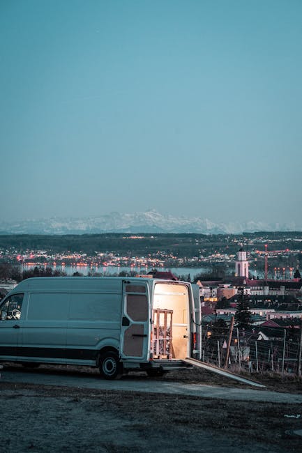A white cargo van is parked on an elevated area with its rear doors wide open, revealing an interior containing a wooden wardrobe and packaging materials. The van is positioned on a paved surface adjacent to a slope, with a ramp leading into the open back door. Nearby, on the ground, there are cardboard boxes and plastic wrapping used for packing household items. In the background, a cityscape with various buildings, residential houses, and industrial structures is visible, along with a scenic view of distant mountains under a clear sky. The scene is illuminated by natural daylight, indicating daytime, and exemplifies a home relocation or furniture transport process as part of house removals services.