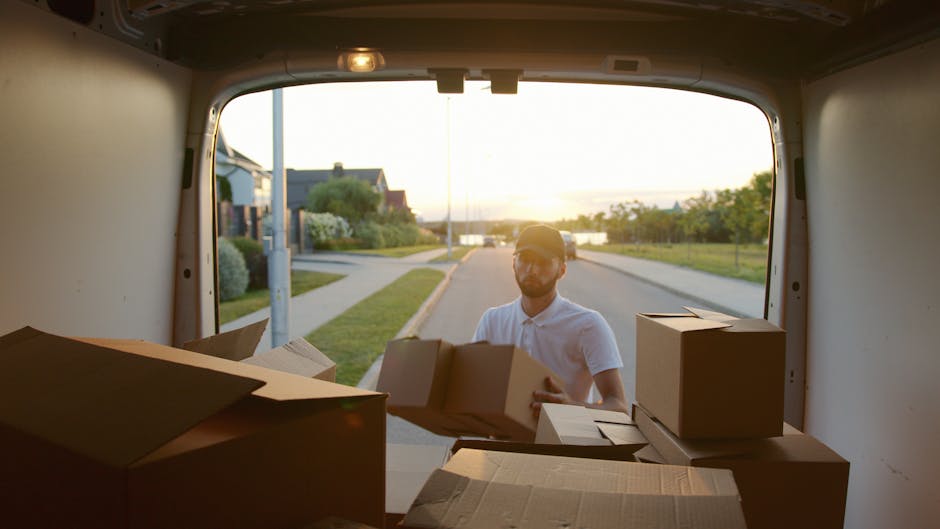 A man wearing glasses and a light-colored polo shirt is seen loading cardboard boxes into the back of a white moving van during a home relocation. The van's interior is visible, showing several sealed cardboard boxes of various sizes, some placed on top of each other. The exterior scene is illuminated by warm, setting sunlight, revealing a suburban street with houses, green lawns, and trees. The man is positioned outside the van, standing on a paved pavement, as he carefully carries a box toward the open rear door of the vehicle, which is engaged in the packing and moving process. This scene reflects professional furniture transport and packing activities typical of house removals, with the focus on the loading phase of a home move facilitated by a removals company like Man with Van Sanderstead, situated within the context of emergency moves in Sanderstead.