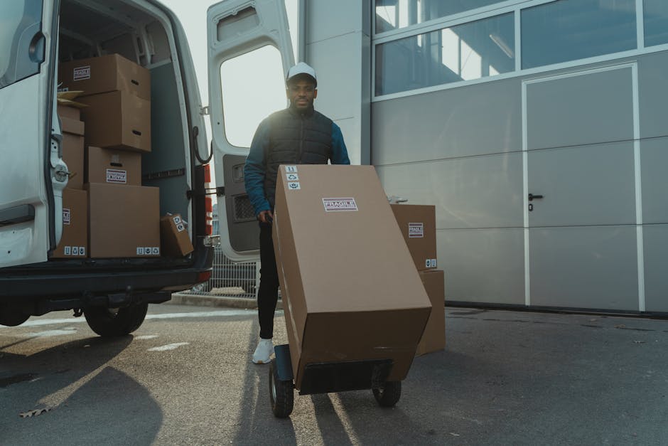 A white cargo van is parked on an elevated area with its rear doors wide open, revealing an interior containing a wooden wardrobe and packaging materials. The van is positioned on a paved surface adjacent to a slope, with a ramp leading into the open back door. Nearby, on the ground, there are cardboard boxes and plastic wrapping used for packing household items. In the background, a cityscape with various buildings, residential houses, and industrial structures is visible, along with a scenic view of distant mountains under a clear sky. The scene is illuminated by natural daylight, indicating daytime, and exemplifies a home relocation or furniture transport process as part of house removals services.