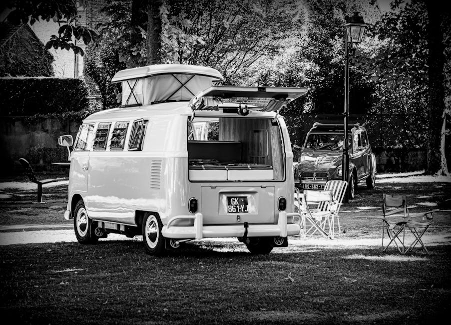 A vintage-style camper van with an open rear hatch revealing a partially visible interior, parked outdoors on a paved area surrounded by trees and foliage. The van's exterior is clean and features a pop-up roof and multiple side windows. Several cardboard boxes, some wrapped in plastic and others unwrapped, are stacked inside the camper, indicating packing for a furniture transport or home relocation. A small utility trailer is positioned behind the van, and a black car is parked nearby, suggesting vehicle transfer or additional moving support. A pole-mounted streetlamp and some outdoor chairs are visible, indicating a small parking or resting area. The scene appears to be in daylight with natural light illuminating the vehicles and surroundings, consistent with a scene of loading or unloading during a house removal or moving process. The presence of Man with Van Sanderstead as the moving service provider subtly supports the context of packing and loading for a house move.