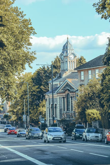 A street scene showing several parked cars along the curb with a large historic clock tower building in the background, surrounded by green leafy trees and residential houses. The building features a tall, ornate clock with Roman numerals, a pointed spire, and decorative architectural details. The sky is clear with some clouds, and the lighting suggests a bright day. The image captures the quiet pavement where a home relocation or furniture transport process could be taking place, with the area well-suited for residential moving services. Man with Van Sanderstead occasionally provides removals, highlighting the importance of efficient packing and loading during house moves within the local community.