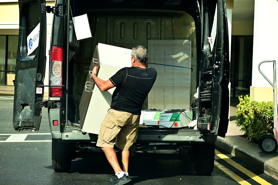 A vintage-style camper van with an open rear hatch revealing a partially visible interior, parked outdoors on a paved area surrounded by trees and foliage. The van's exterior is clean and features a pop-up roof and multiple side windows. Several cardboard boxes, some wrapped in plastic and others unwrapped, are stacked inside the camper, indicating packing for a furniture transport or home relocation. A small utility trailer is positioned behind the van, and a black car is parked nearby, suggesting vehicle transfer or additional moving support. A pole-mounted streetlamp and some outdoor chairs are visible, indicating a small parking or resting area. The scene appears to be in daylight with natural light illuminating the vehicles and surroundings, consistent with a scene of loading or unloading during a house removal or moving process. The presence of Man with Van Sanderstead as the moving service provider subtly supports the context of packing and loading for a house move.