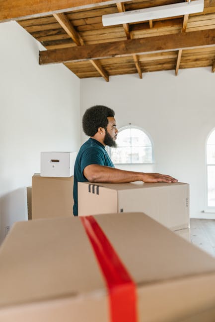 A man with dark curly hair and a beard, wearing a blue T-shirt, is standing inside a room with white walls and a wooden ceiling, preparing for a home relocation. He is positioned behind a large cardboard box, resting his hands on top of it, with additional boxes stacked nearby. The boxes are sealed with packing tape, some wrapped with red straps for secure lifting. The room features two large arched windows allowing natural light to illuminate the space, which has been cleared of furniture to facilitate the furniture transport process. The environment appears to be part of the initial packing stage or loading area for a professional removal service, such as those offered by Man with Van Sanderstead. The setting emphasizes careful handling and organization during packing and moving activities typical of house removals.