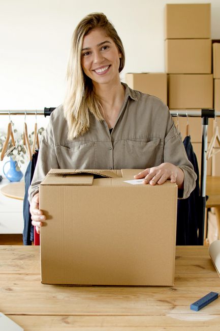 A young woman with long blonde hair and a friendly smile, wearing a light grey shirt, stands indoors holding a medium-sized cardboard box with her left hand resting on top. Behind her, there is a clot