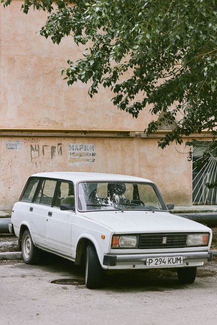 A vintage white station wagon vehicle is parked on a street beside a beige wall with some graffiti and a metal gate. The car has a boxy design typical of older models, featuring rectangular headlights