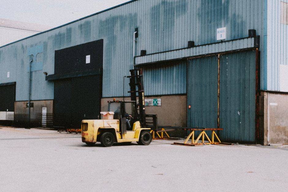 A yellow forklift truck is positioned outside a large industrial warehouse with blue corrugated metal walls, preparing to load or unload items. The forklift's forks are lowered and aligned with a set of orange safety barriers on the pavement directly in front of a wide sliding metal door, which is part of the warehouse's outdoor loading area. The warehouse features another large black loading bay door to the left with a small elevated platform nearby, and a white sign above the main door displaying steps for safety procedures. The area is well-lit with natural daylight, with no other vehicles or personnel visible in the immediate vicinity. This scene captures the logistical process involved in home relocation or furniture transport, showcasing the equipment and environment typical for professional removals services like those offered by Man with Van Sanderstead, supporting efficient packing and moving operations.