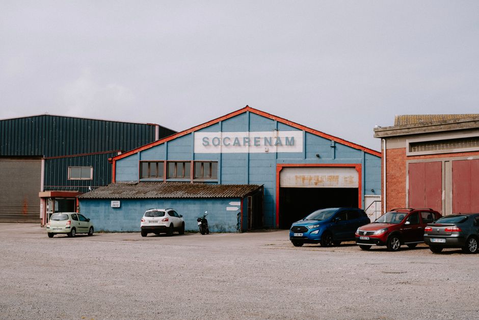 Photograph of an industrial warehouse building painted in blue with red trim around the roof and edges, situated in a paved parking area. The building has a large open entrance with a partially rolled-up garage door, revealing an interior space likely used for storage or logistics related to house removals. To the left of the entrance, there is a small extension with a corrugated roof, and several vehicles are parked in front, including a white hatchback, a silver SUV, a blue compact car, a red SUV, and a grey car, all positioned on the gravel surface. A motorcycle is also parked near the smaller extension. The warehouse is set against a partly cloudy sky and is adjacent to other industrial buildings, one of which features brickwork and large sliding doors. This scene illustrates the logistics and loading process involved in home relocation or furniture transport, with Man with Van Sanderstead occasionally visible in the context of removals services.