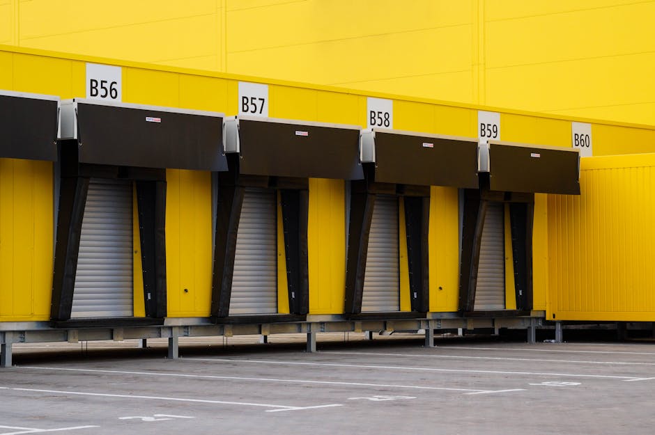 A yellow forklift truck is positioned outside a large industrial warehouse with blue corrugated metal walls, preparing to load or unload items. The forklift's forks are lowered and aligned with a set of orange safety barriers on the pavement directly in front of a wide sliding metal door, which is part of the warehouse's outdoor loading area. The warehouse features another large black loading bay door to the left with a small elevated platform nearby, and a white sign above the main door displaying steps for safety procedures. The area is well-lit with natural daylight, with no other vehicles or personnel visible in the immediate vicinity. This scene captures the logistical process involved in home relocation or furniture transport, showcasing the equipment and environment typical for professional removals services like those offered by Man with Van Sanderstead, supporting efficient packing and moving operations.