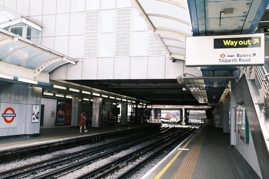 Image of the platform at Hammersmith underground station with multiple train tracks and a raised walkway above, featuring white and grey tiled walls and a curved, transparent roof covering the station area. On the right side, there is a staircase with metal railings leading to an upper level, alongside an overhead sign indicating the 'Way out' towards Buses and Talgarth Road. The platform appears to be part of a busy urban commute setting, with a few passengers visible on the left side, dressed in casual clothing. In the distance, a train can be seen approaching or departing along the tracks. This environment reflects the typical scene at a London underground station, relevant to house removals or moving logistics when coordinating transport to or from the station, as handled by companies like Man with Van Sanderstead.