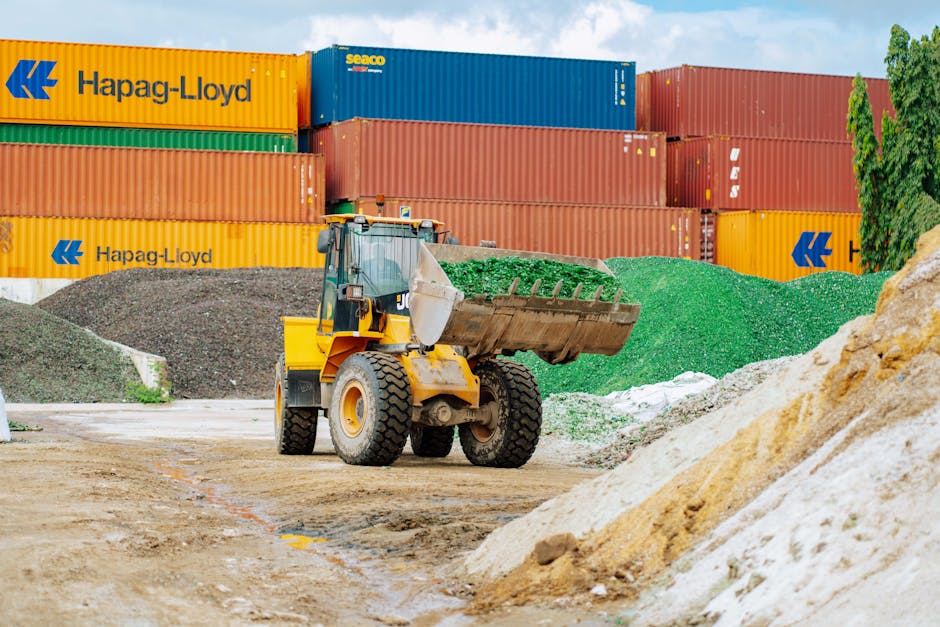 A construction or moving site featuring a yellow front loader with a large bucket full of green gravel or crushed stone, positioned on a dirt ground. Behind the loader, there are multiple large shipping containers stacked horizontally, painted in bright yellow, blue, and red, with some bearing the 'Hapag-Lloyd' logo. To the right, there is a pile of light brown sand and smaller piles of green and white aggregate materials. The scene is outdoors, under natural daylight, with a backdrop of green trees and a partly cloudy sky. The limited surrounding space and narrow roads suggest a challenge in household or furniture transport during a home relocation process. The setting highlights the logistical considerations of moving furniture and boxes, with the loader and containers indicative of construction, renovation, or cargo transport related to moving services, exemplified by [COMPANY_NAME].