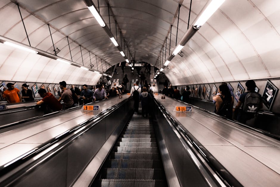 Image of the platform at Hammersmith underground station with multiple train tracks and a raised walkway above, featuring white and grey tiled walls and a curved, transparent roof covering the station area. On the right side, there is a staircase with metal railings leading to an upper level, alongside an overhead sign indicating the 'Way out' towards Buses and Talgarth Road. The platform appears to be part of a busy urban commute setting, with a few passengers visible on the left side, dressed in casual clothing. In the distance, a train can be seen approaching or departing along the tracks. This environment reflects the typical scene at a London underground station, relevant to house removals or moving logistics when coordinating transport to or from the station, as handled by companies like Man with Van Sanderstead.