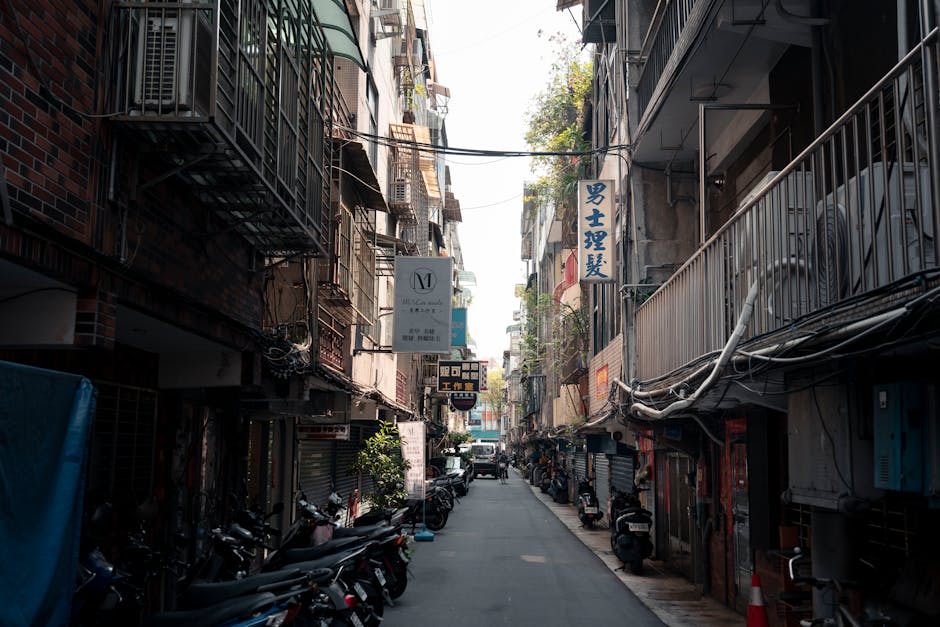 A narrow residential alleyway with brick and concrete exterior walls on either side, leading to a small, well-maintained street with houses featuring red-tiled roofs and front gardens. A woman with sh