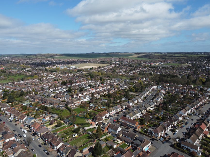 Aerial view of a suburban neighbourhood with rows of terraced houses and semi-detached homes, featuring visible back gardens and tree-lined streets. In the background, open fields and rolling countryside extend to the horizon under a partly cloudy sky. The image highlights a typical residential area, possibly during daylight hours with natural lighting. This setting may relate to house relocations or moving services provided by Man with Van Sanderstead, illustrating the environment where furniture transport and packing and moving activities could take place during a south Croydon estate removal.