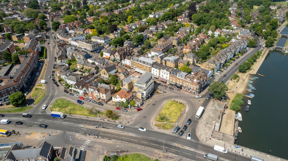 An aerial view of South Croydon near Sanderstead, showing a town centre with a mix of terraced and detached houses, retail shops, and a parking area. The scene includes a large river or lake on the right side with boats docked along the shoreline and a paved promenade. Several vehicles such as cars, vans, and trucks are parked along the streets and in the parking areas, with some engaged in loading or unloading activities. The surrounding environment features tree-lined streets, green spaces, and residential buildings with pitched roofs. The image captures the movement and urban layout typical of a community undergoing home relocation or furniture transport, illustrating the context in which Man with Van Sanderstead’s removals services might be involved in packing, loading, or transit processes.