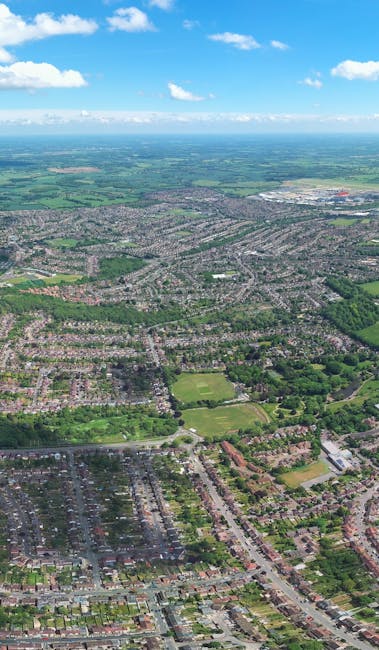 Aerial view of a suburban neighbourhood with rows of terraced houses and semi-detached homes, featuring visible back gardens and tree-lined streets. In the background, open fields and rolling countryside extend to the horizon under a partly cloudy sky. The image highlights a typical residential area, possibly during daylight hours with natural lighting. This setting may relate to house relocations or moving services provided by Man with Van Sanderstead, illustrating the environment where furniture transport and packing and moving activities could take place during a south Croydon estate removal.