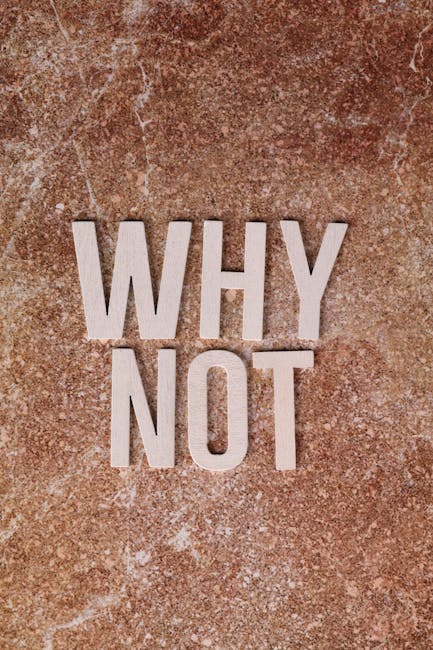 Close-up of a textured, reddish-brown stone wall with the words 'WHY NOT' spelled out in large, light-colored wooden or stone letters arranged vertically. The surface shows natural variations and speckles, providing a neutral background. This image may be used to illustrate topics related to decision-making or prompting reflection. It does not depict any furniture, boxes, or moving equipment, but could be contextually associated with house removals or relocation services, emphasizing questions about choices or planning processes involved in home relocations. The image's lighting highlights the rough texture of the wall and the contrast between the background and the lettering, suitable for use on a webpage discussing transparent pricing or the reasoning behind quotation variances in house removals services, such as those provided by Man with Van Sanderstead.